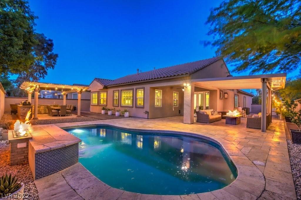 A beautiful, desert style home in the evening featuring two covered patio spaces, well lit with a pool, seating area, and mature trees.